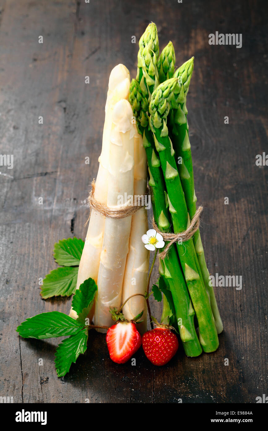 Upright bunches of fresh green and white asparagus spears balanced on a wooden table top garnished with strawberries Stock Photo