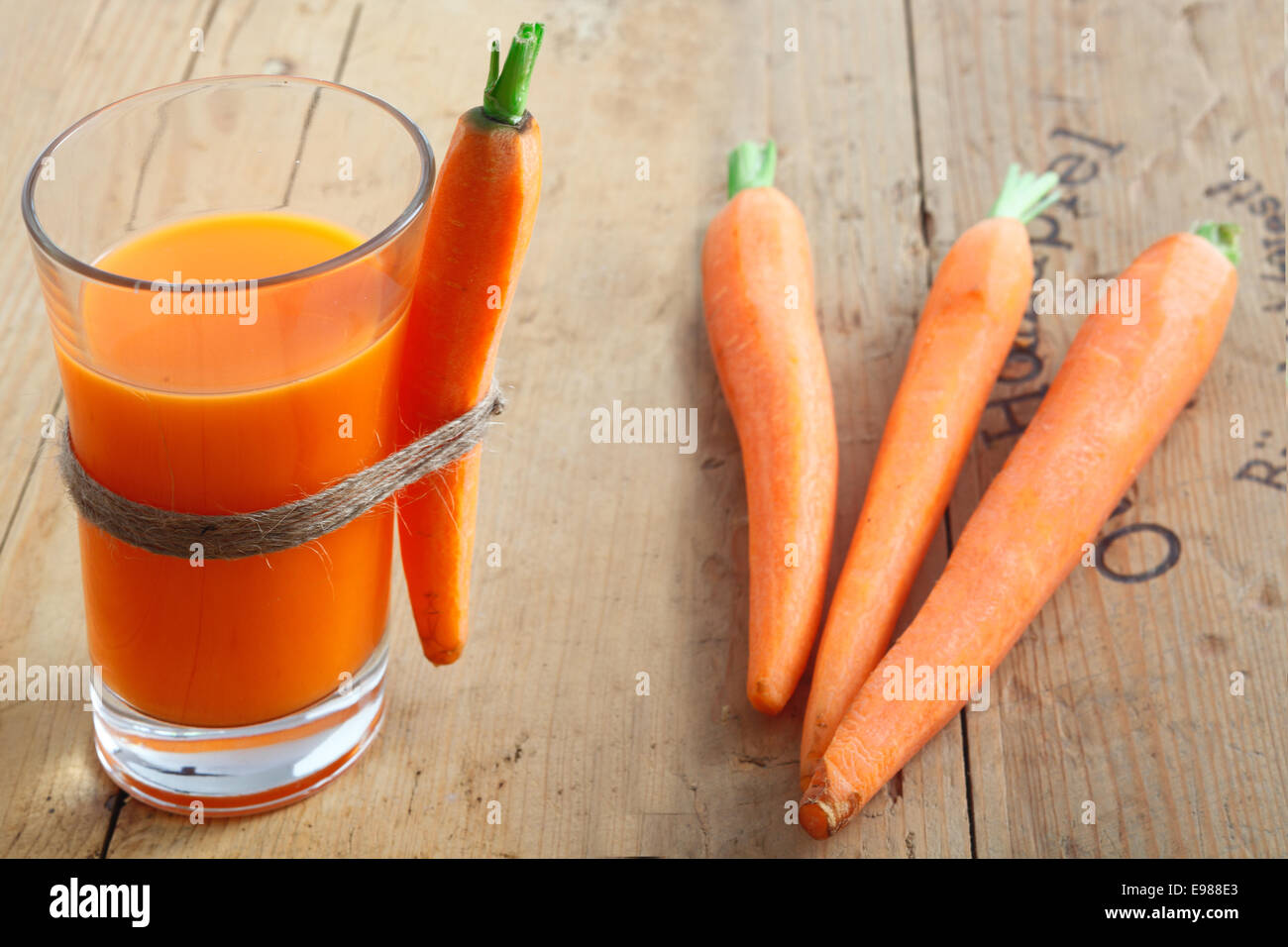 A tall glass of healthy freshly blended carrot juice on a wooden table with a whole carrot attached and a trio of raw carrots lying alongside Stock Photo