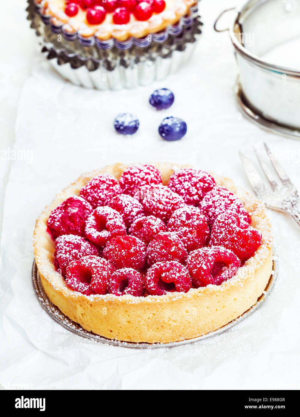 Small individual serving of a fresh whole raspberry tart dusted with sugar on a golden crisp crust with a silver cake fork Stock Photo