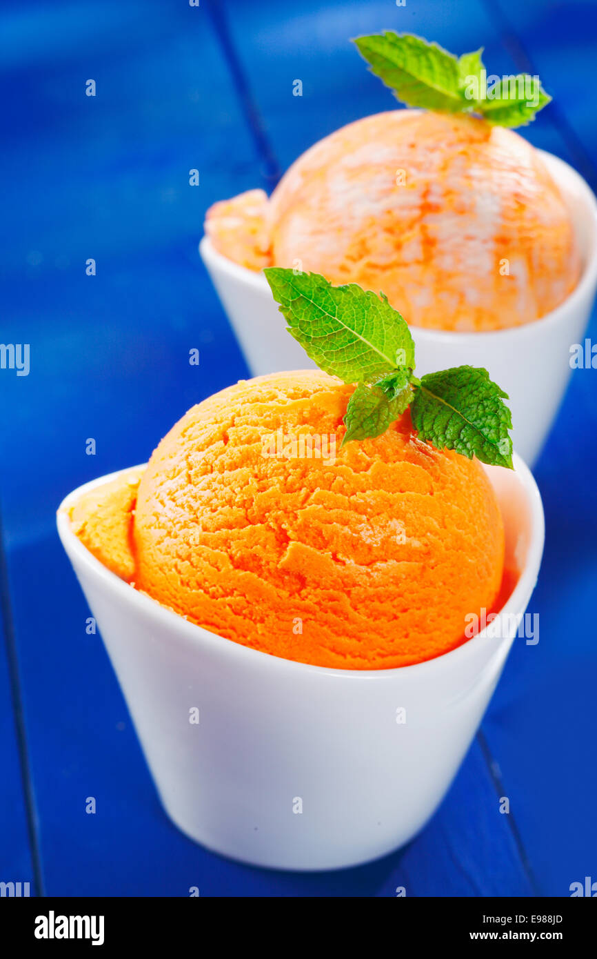 Closeup of a round scoop of colourful creamy orange icecream garnished with mint in boat-shaped porcelain bowls on blue boards Stock Photo