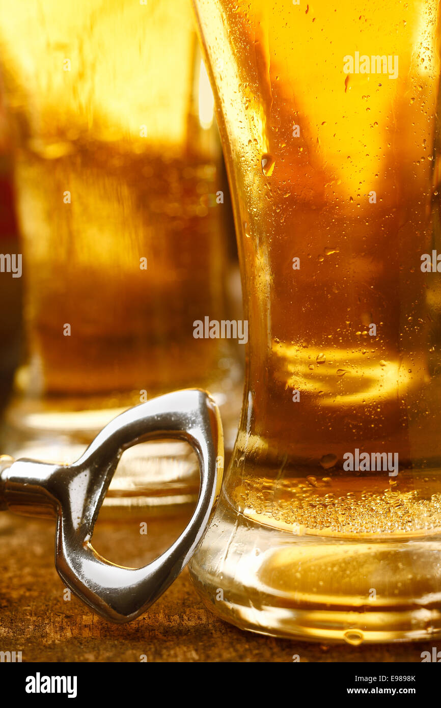 Bottle opener on a wooden tabletop leaning against a tall glass full of chilled golden beer, closeup cropped image Stock Photo
