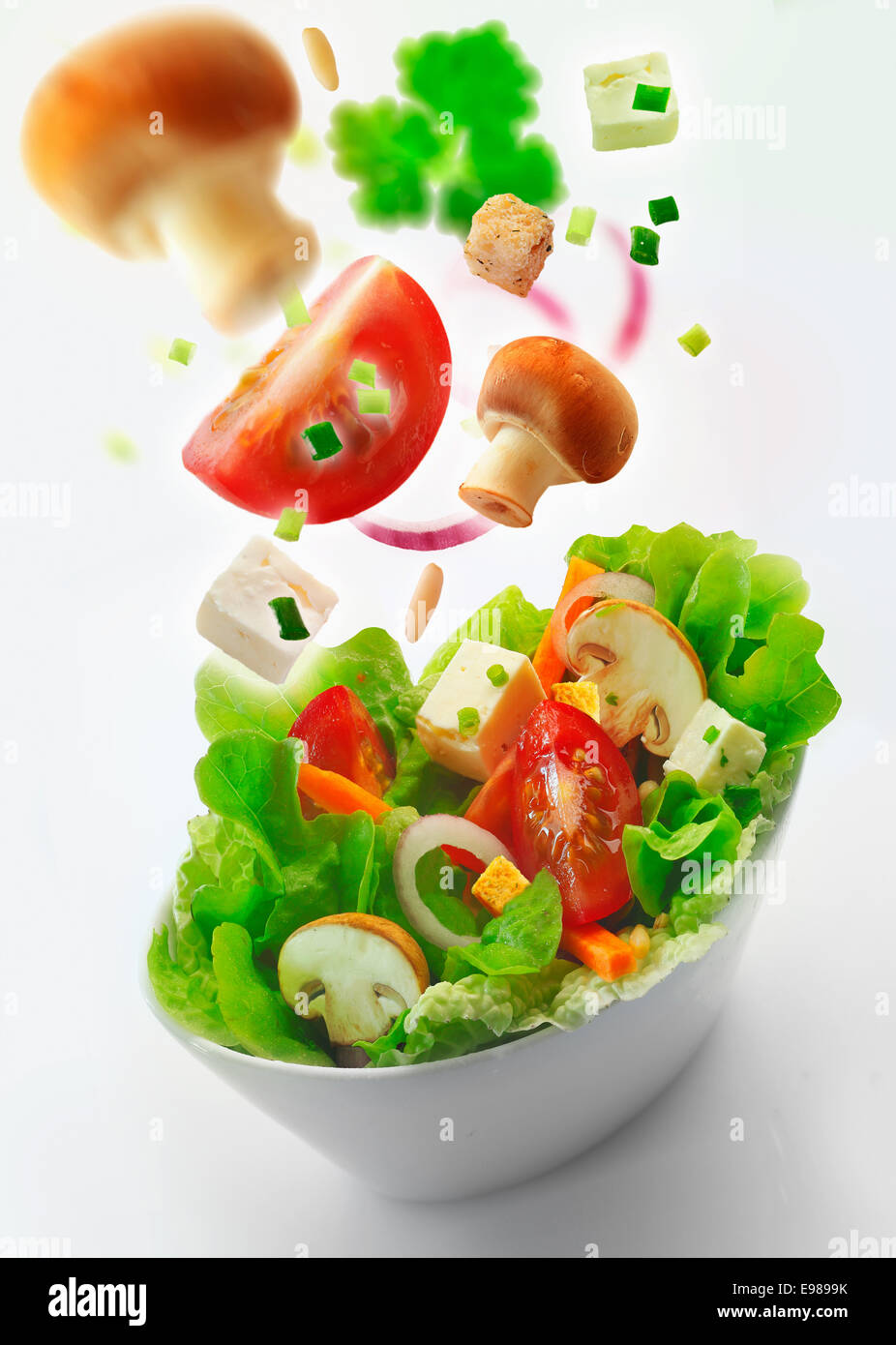 Individual side bowl of healthy fresh mixed green salad of lettuce, carrot, onion, mushrooms, tomato and feta with additional ingredients in th air receding into the distance on a white background Stock Photo