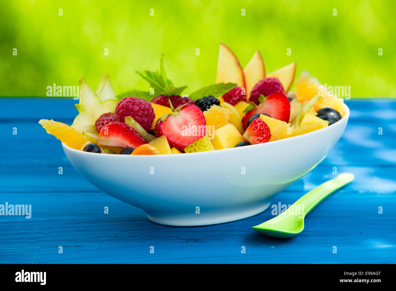 Mix of pieces of fresh delicious fruits in a bowl, on a table against a blurred green background Stock Photo