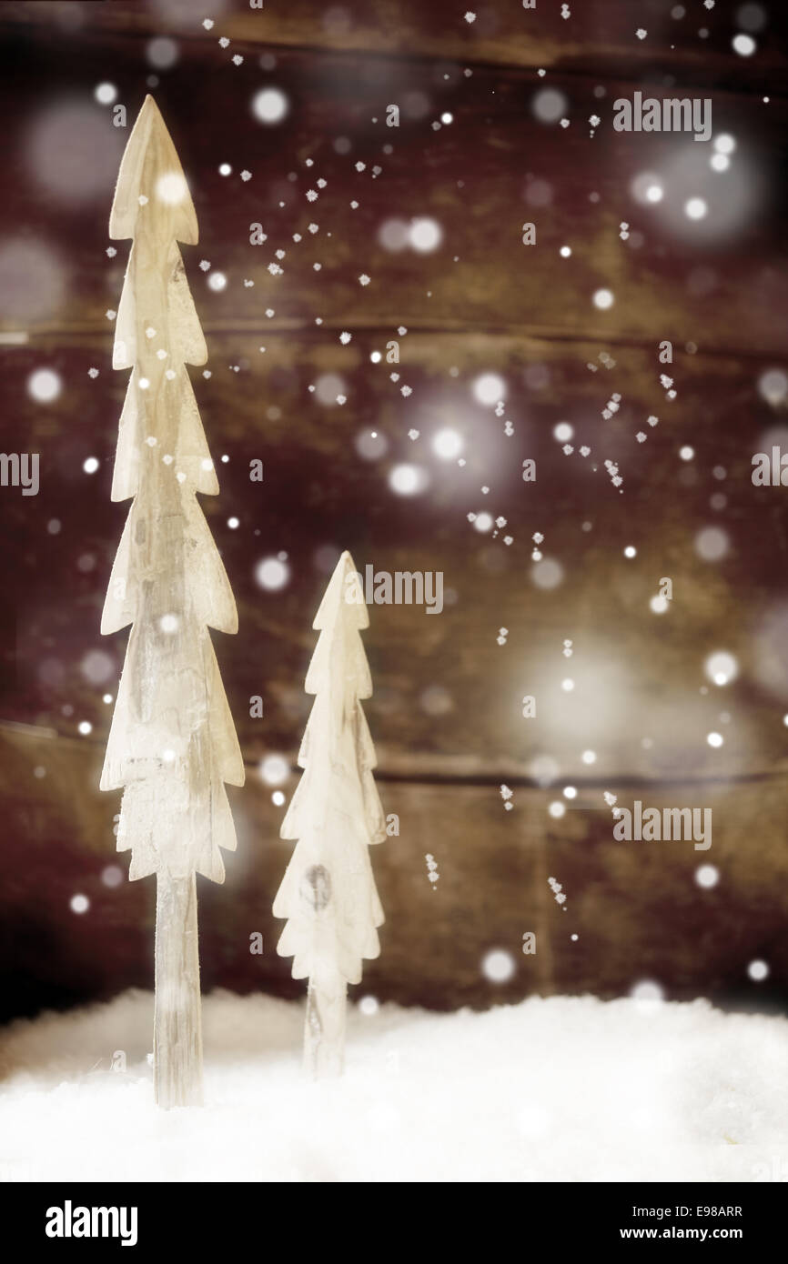 Two simple rustic Christmas trees cut out of wood standing in snow in front of a wooden wall with falling snowflakes for a festive Christmas winter background Stock Photo