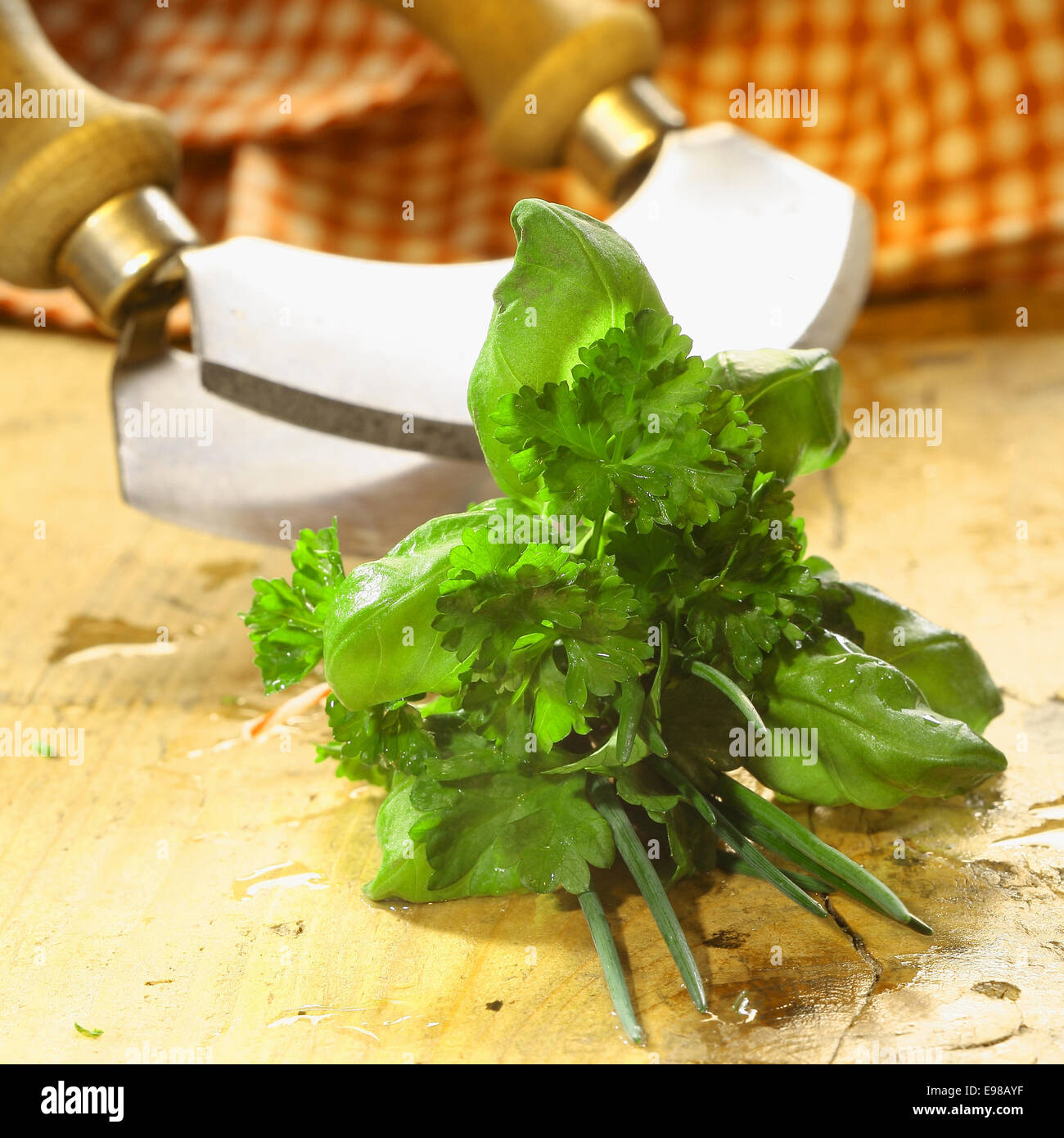 Bouquet garni of fresh herbs including basil, crinkly leaf parsley and chives lying on a wooden table in front of a chopping blade Stock Photo