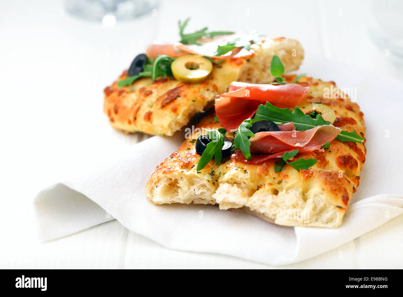 Fresh oven-baked golden focaccia bread with ham, sliced olives and fresh rocket leaves served on a white napkin Stock Photo