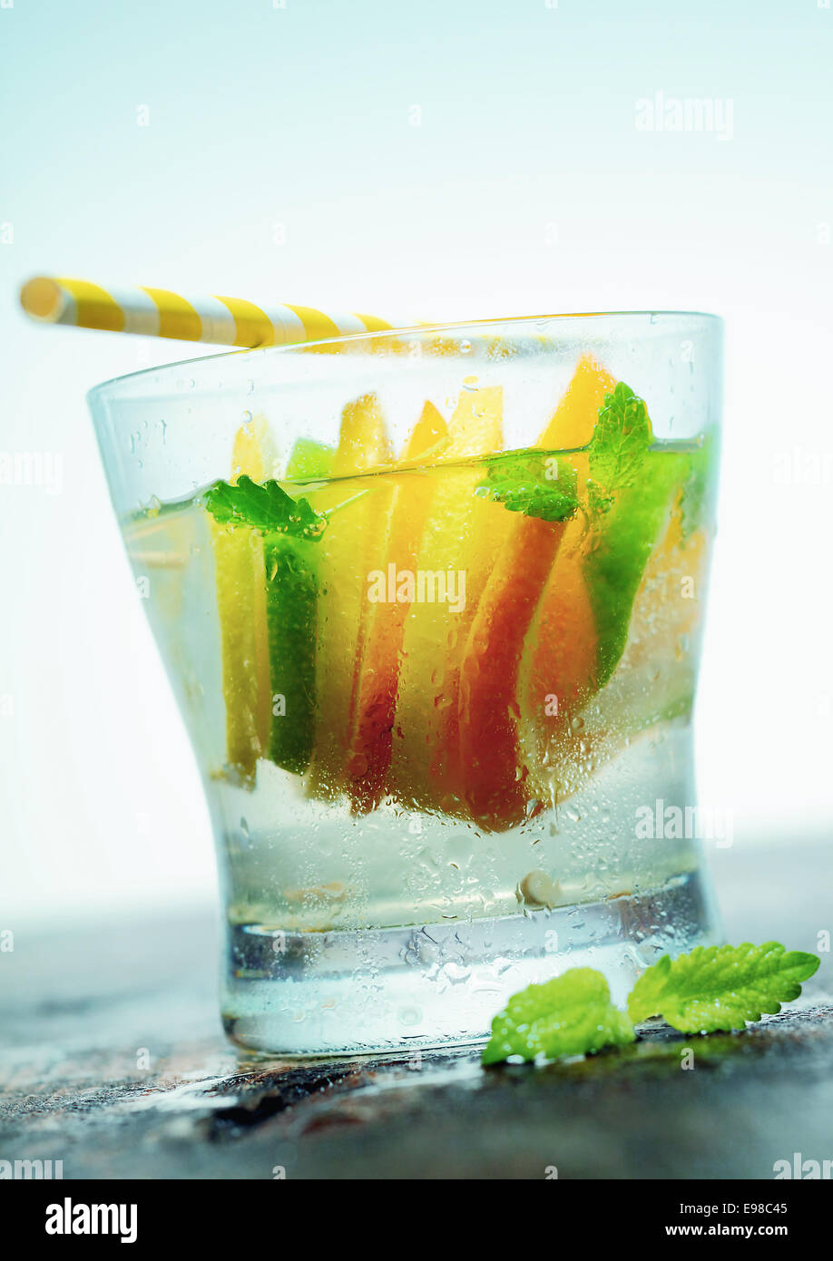 Glass of refreshing iced vodka or gin with slices of tangy citrus and fresh leaves of mint served on an old wooden countertop with a straw Stock Photo