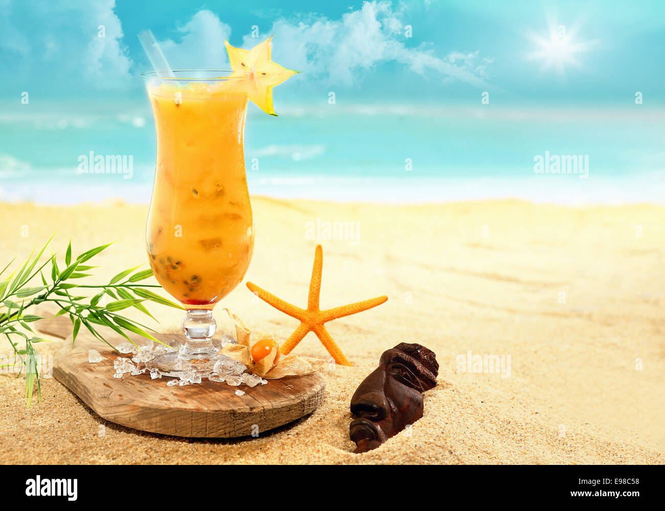 Colourful carambola and orange cocktail ina tall glass served on a wooden board on a golden beach at a tropical holiday resort during an enjoyable summer vacation Stock Photo