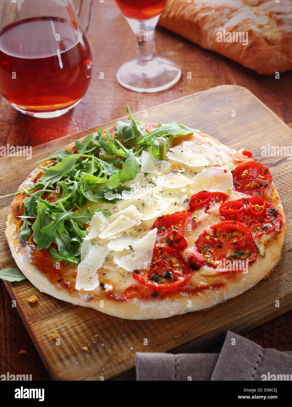 Italian pizza in the red, white and green colours of the national flag formed by the three toppings of tomatoes, cheese shavings and fresh rocket leaves on a wooden board served with a light red wine Stock Photo