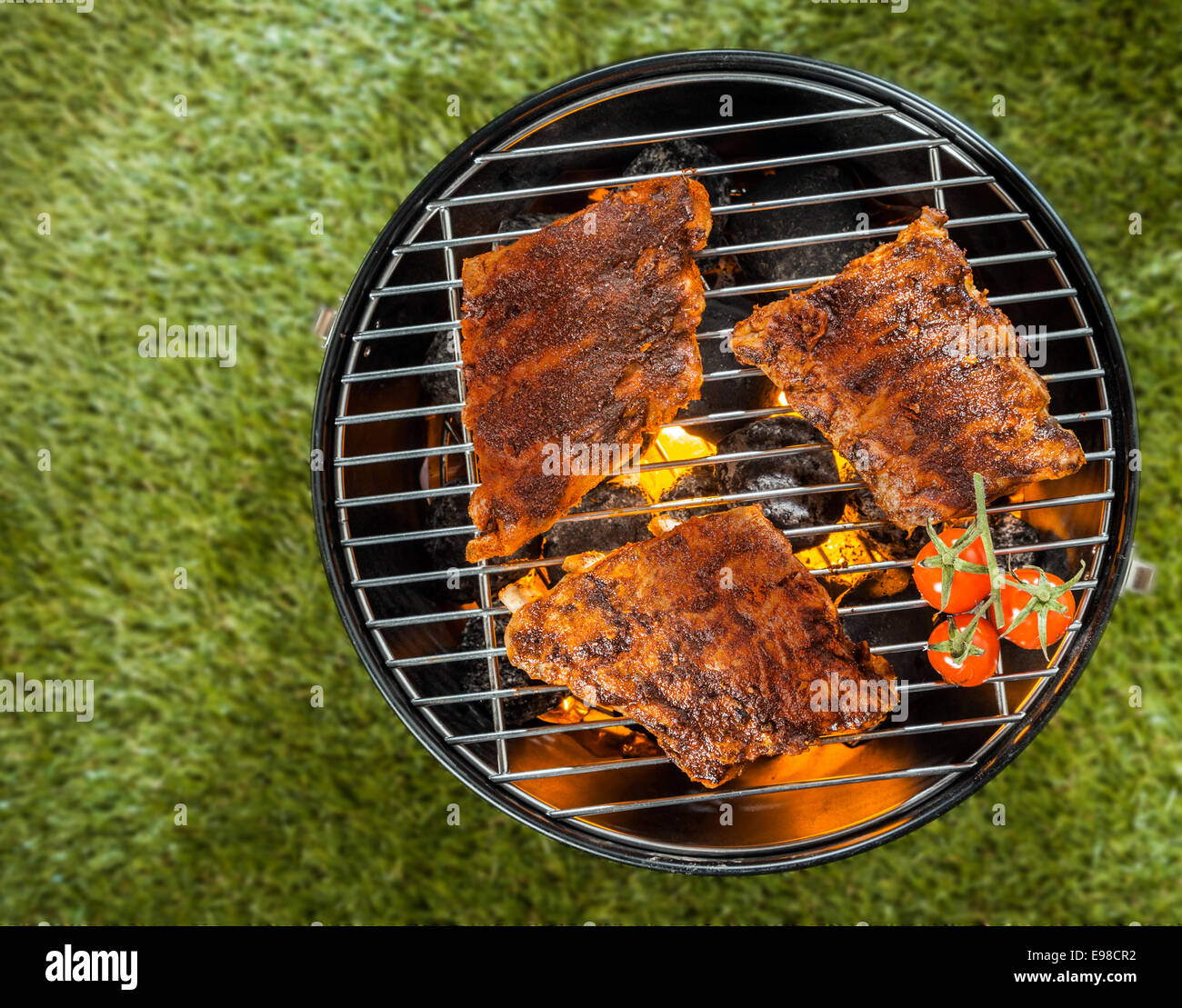 Overhead view of three portions of spicy marinated ribs grilling on a portable metal BBQ on green grass at a summer picnic with Stock Photo