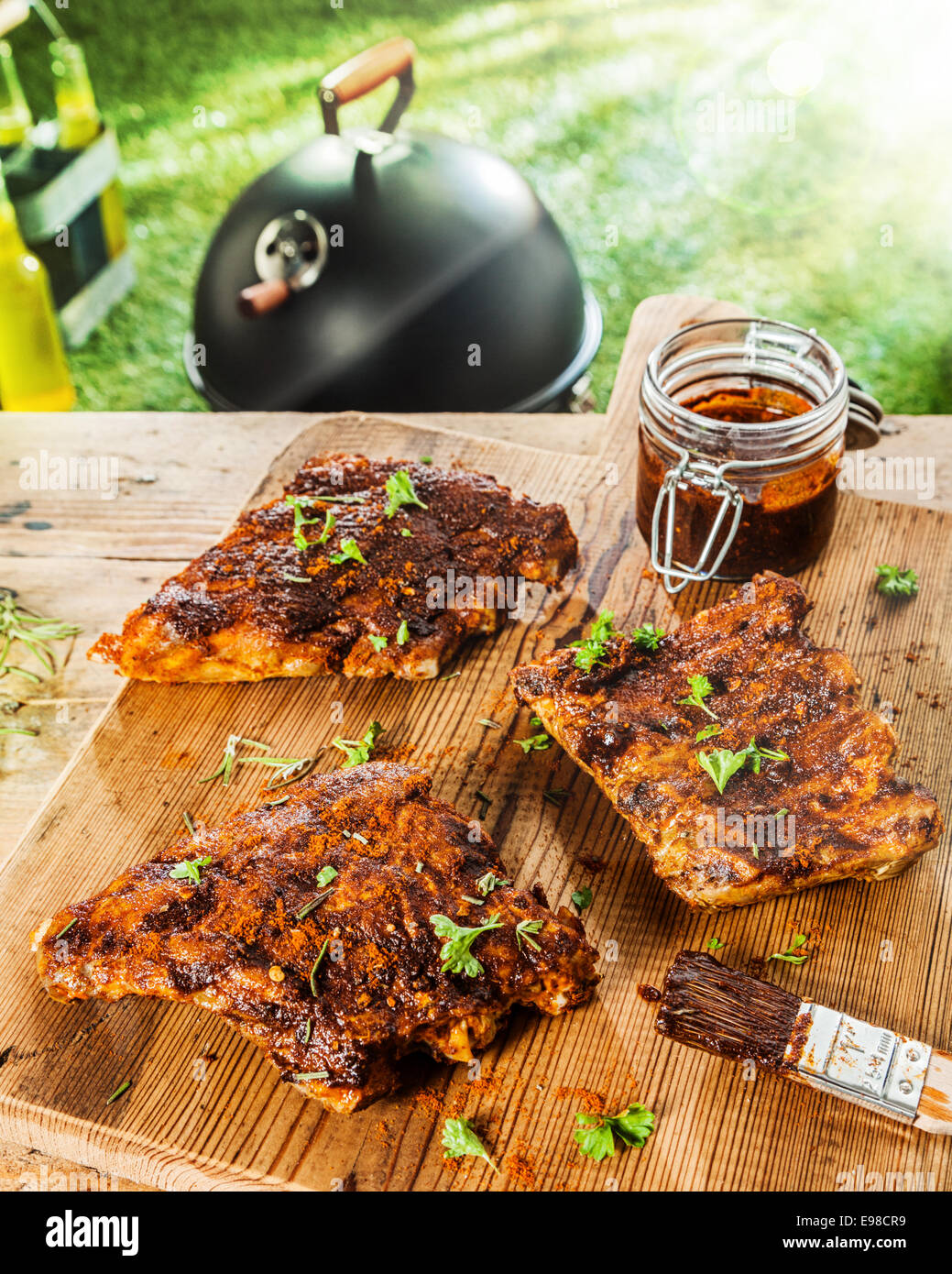 Preparing ribs for a BBQ with savory basting sauce and finely chopped fresh herbs with three portions on a table with a jar of marinade and a brush Stock Photo