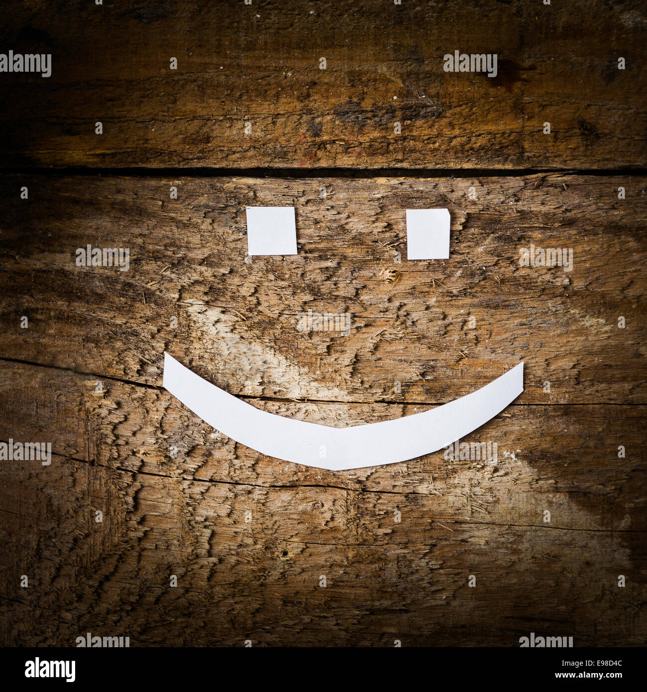 Happy smiling face emoticon from cutout white paper on a grungy rough weathered timber plank with wood grain detail and side vignetting Stock Photo