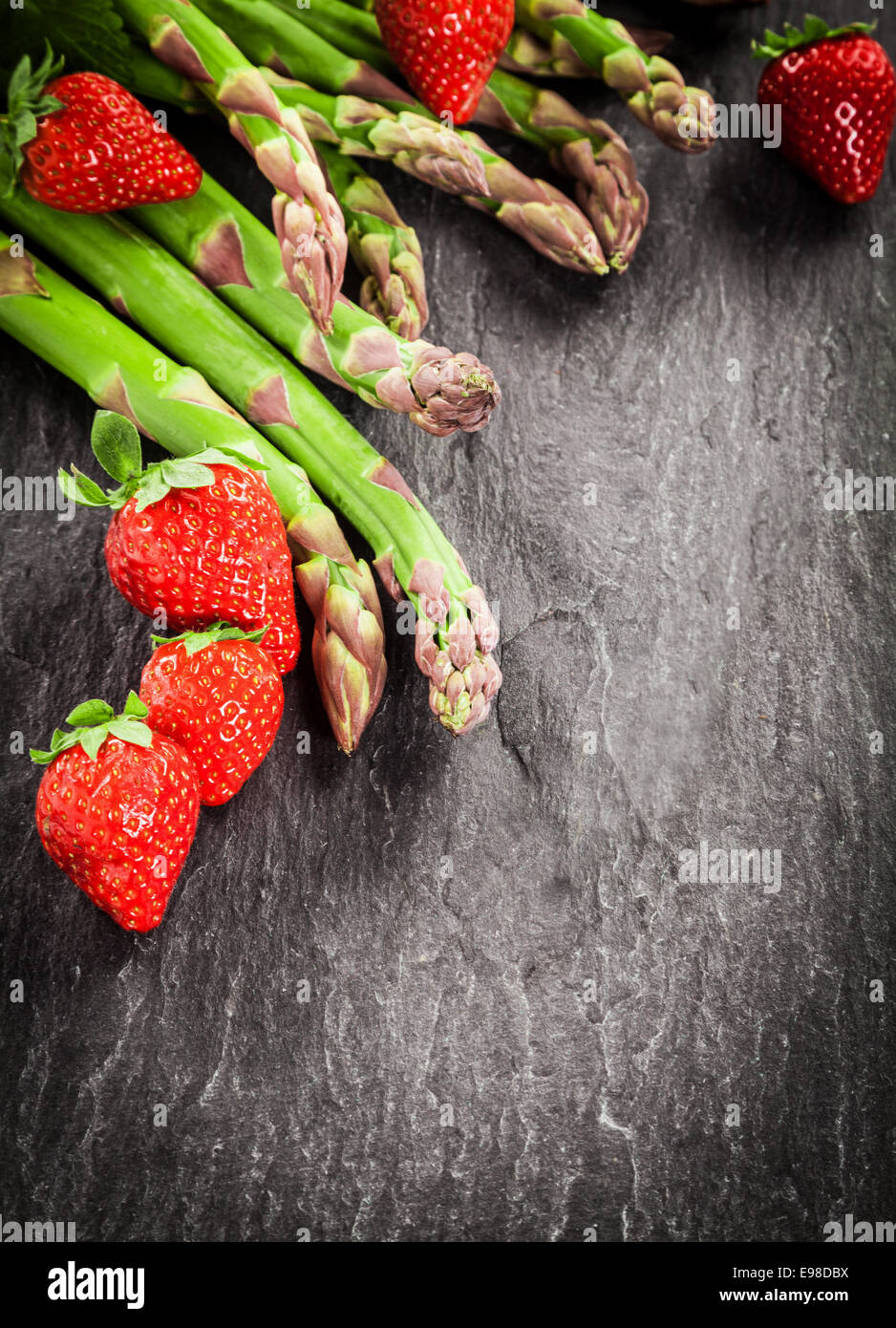 Fresh uncooked green asparagus spears and ripe red strawberries arranged as a corner decoration on textured grey slate with copyspace and vignetting Stock Photo
