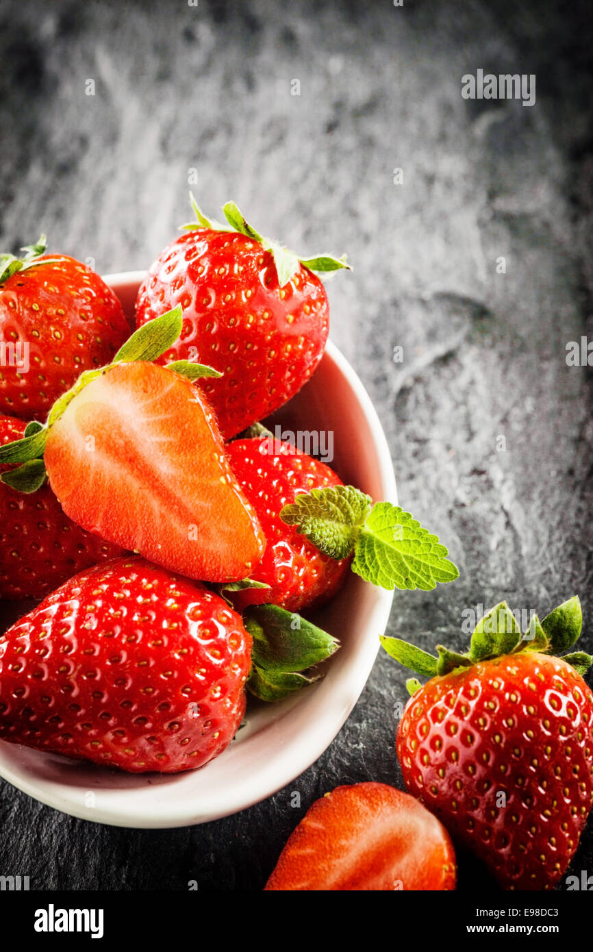 Bowl of ripe red strawberries with green stalks and one halved to show the juicy pulp, high angle view on a textured grey slate Stock Photo