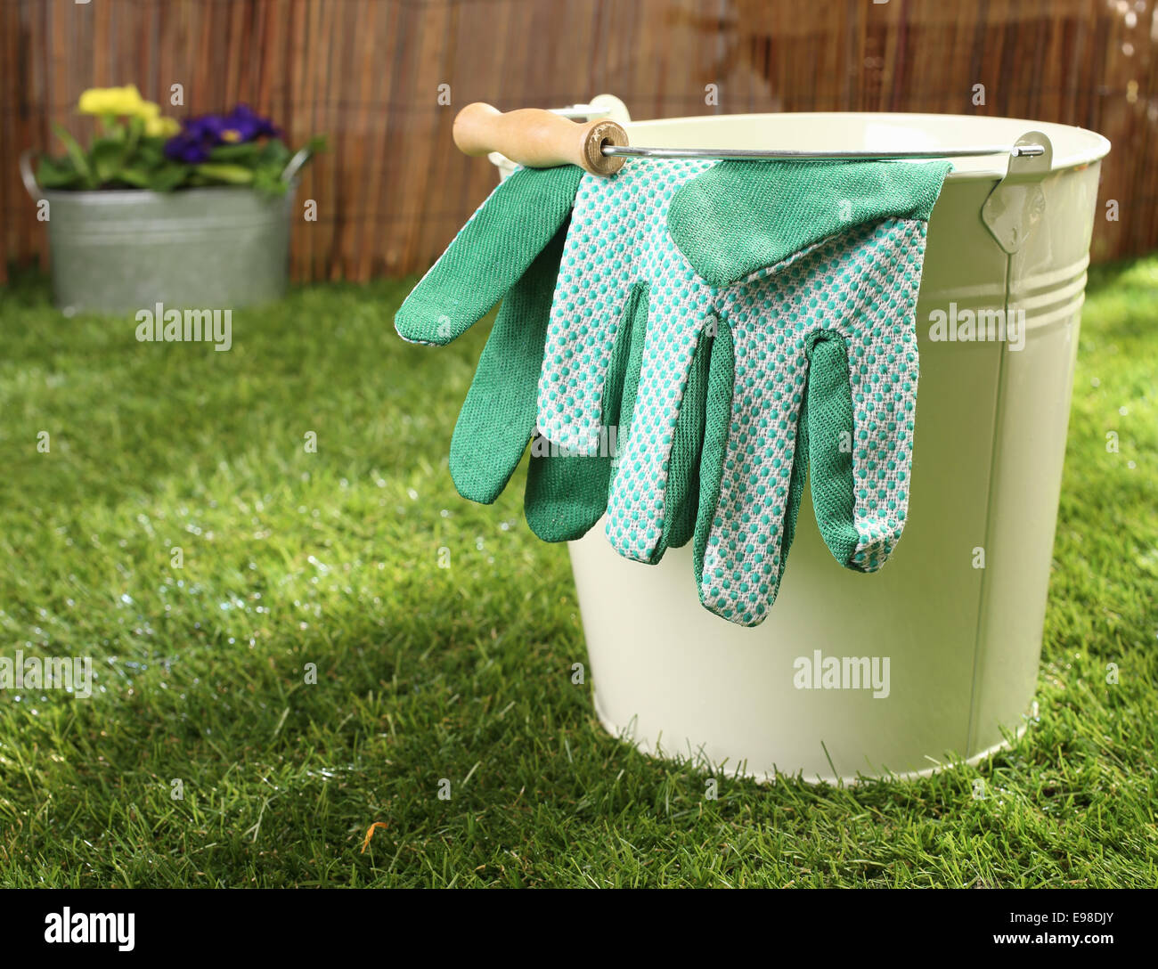 Green textile gardening gloves hanging over the rim of a metal bucket standing on a green lawn in summer sunshine, conceptual image Stock Photo