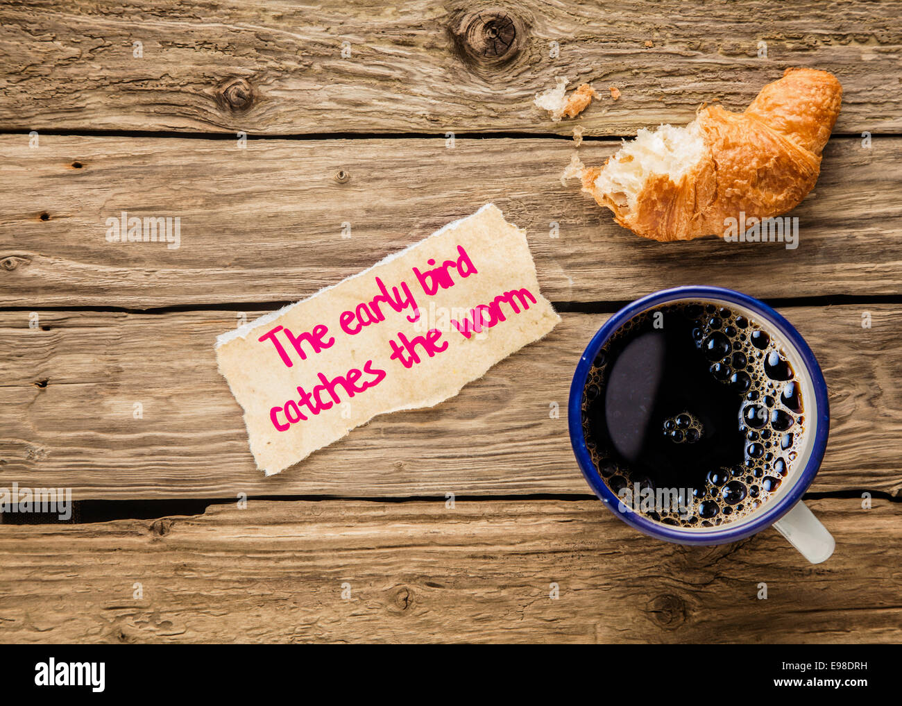 The early bird catches the worm, an inspirational saying hand written on a small torn piece of paper alongside an early breakfast of frothy espresso coffee and a half devoured fresh golden croissant Stock Photo