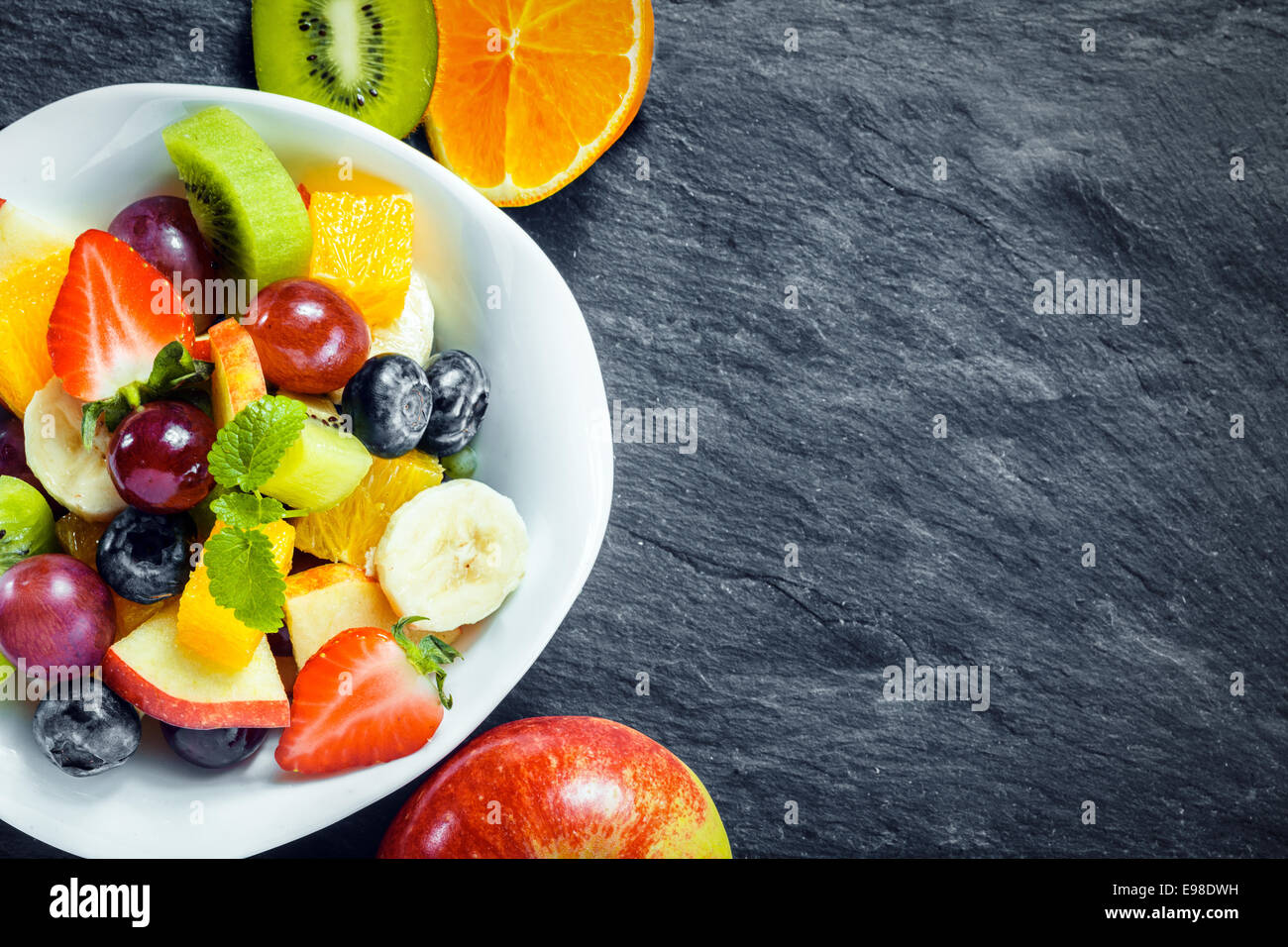 Refreshing bowl of fresh tropical fruit salad with ingredients on a textured slate kitchen counter with copyspace, overhead view Stock Photo