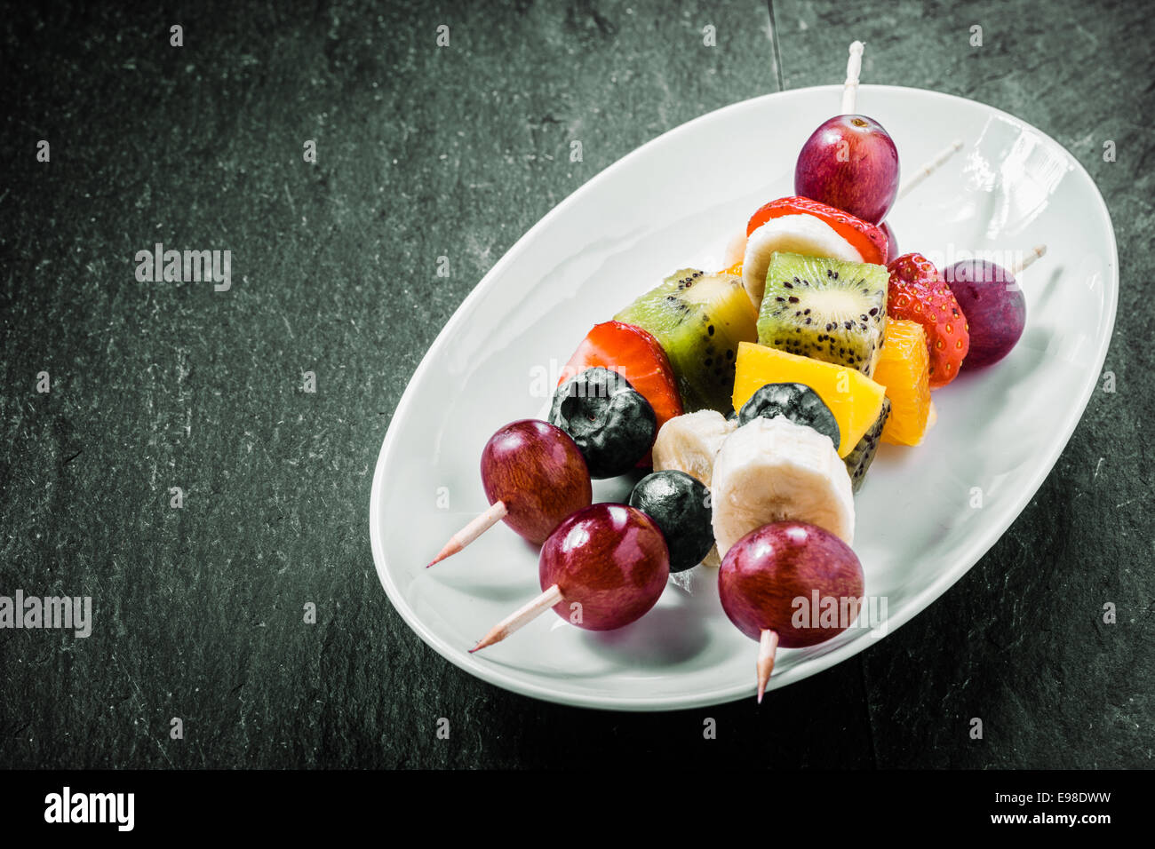 Colorful exotic fruit kebabs served on an oval dish with grapes, kiwifruit, orange, banana, blueberries, and strawberries on a dark background with copyspace Stock Photo