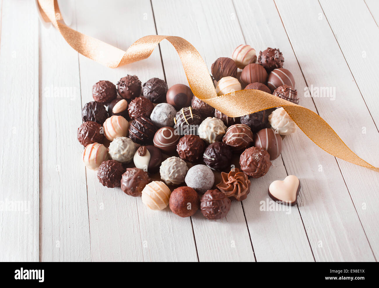 Heart shape made with various types of chocolate truffles decorated with a golden ribbon over a white wooden table Stock Photo