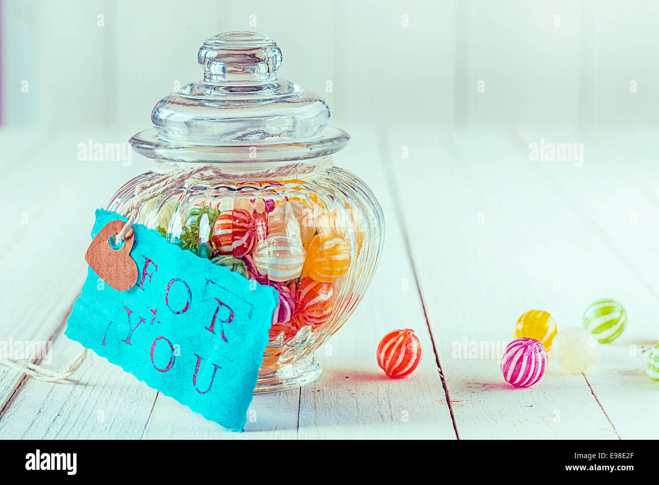 Close-up view of an antique candy jar filled with colorful candies with a blue tag with a romantic, For You, written on a wooden background Stock Photo