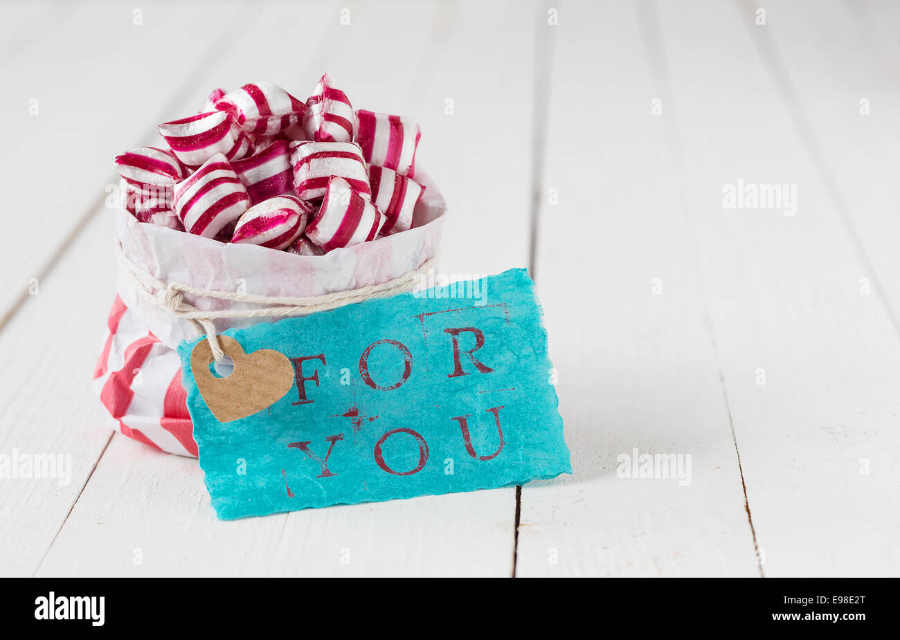 Gift of red and white striped candy in a paper packet with a blue tag with the words For You on a background of white wooden boards Stock Photo