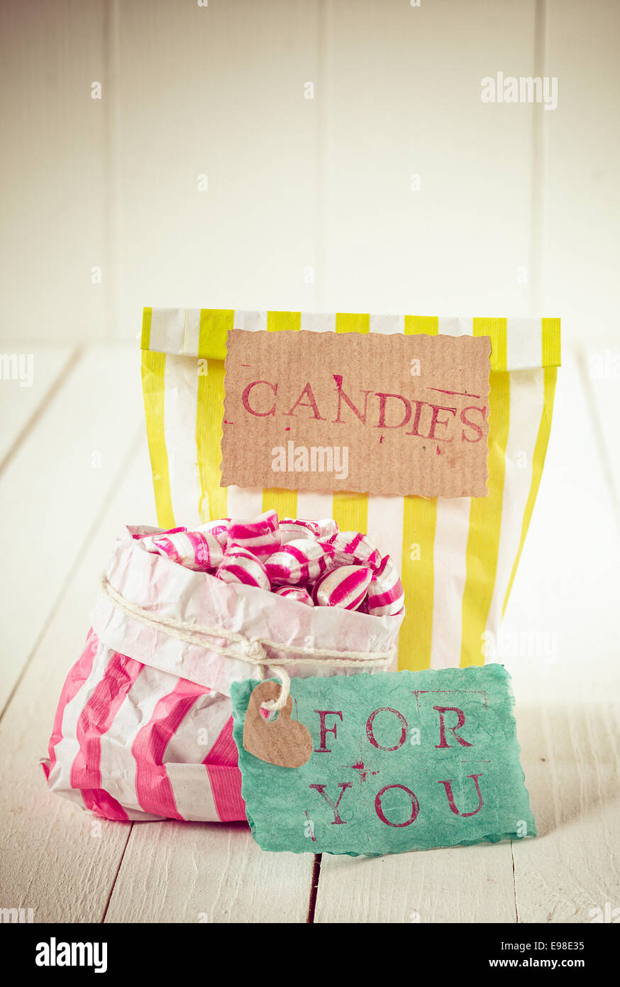 Two colorful bags with white, yellow and pink stripes filled with candies with romantic message in tags meaning, Candies For You, over a white wooden background Stock Photo