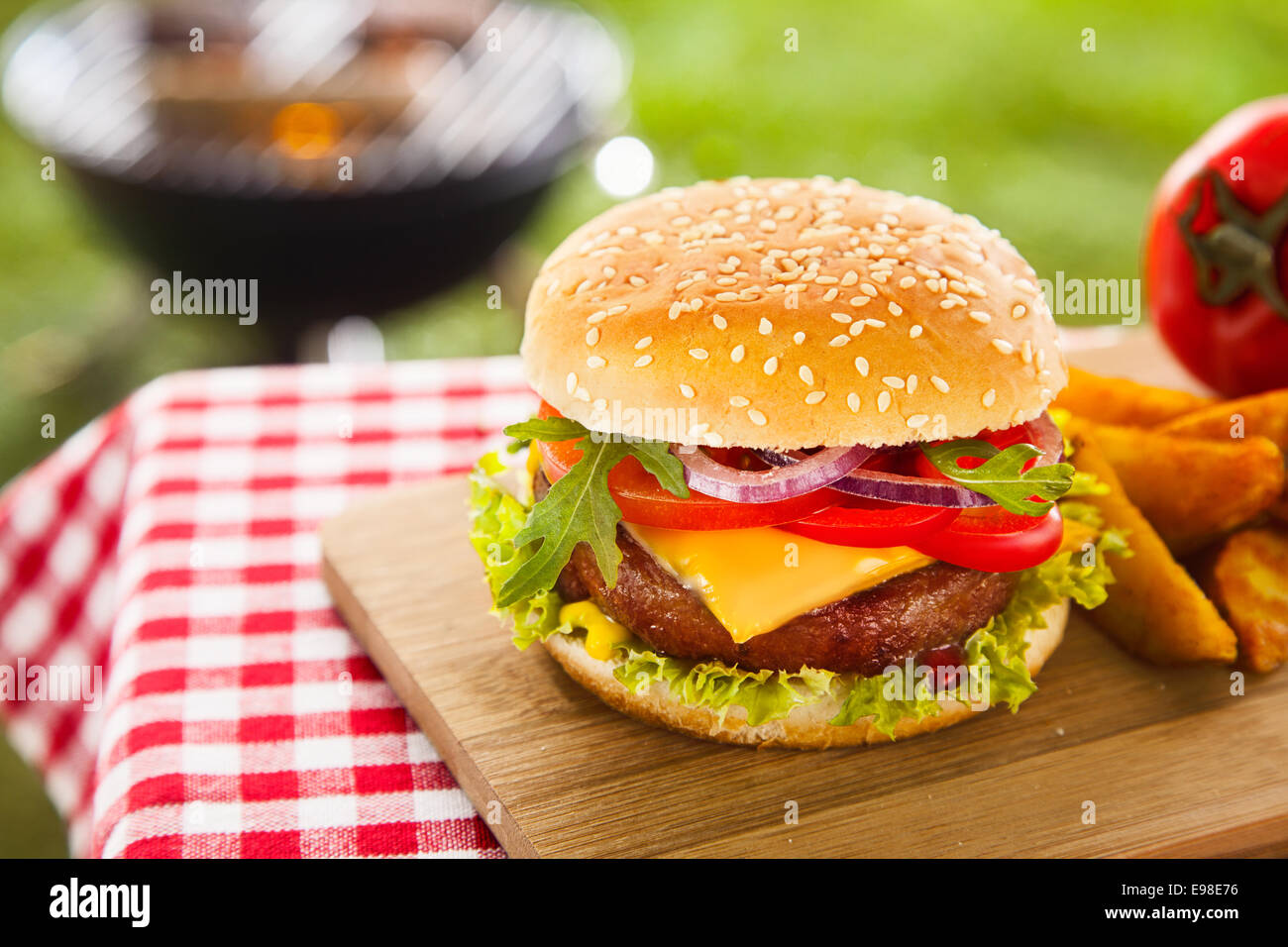 Tasty cheeseburger with melted cheddar cheese dripping over ground beef burger garnished with fresh salad ingredients and served on a wooden table on an outdoor picnic table Stock Photo