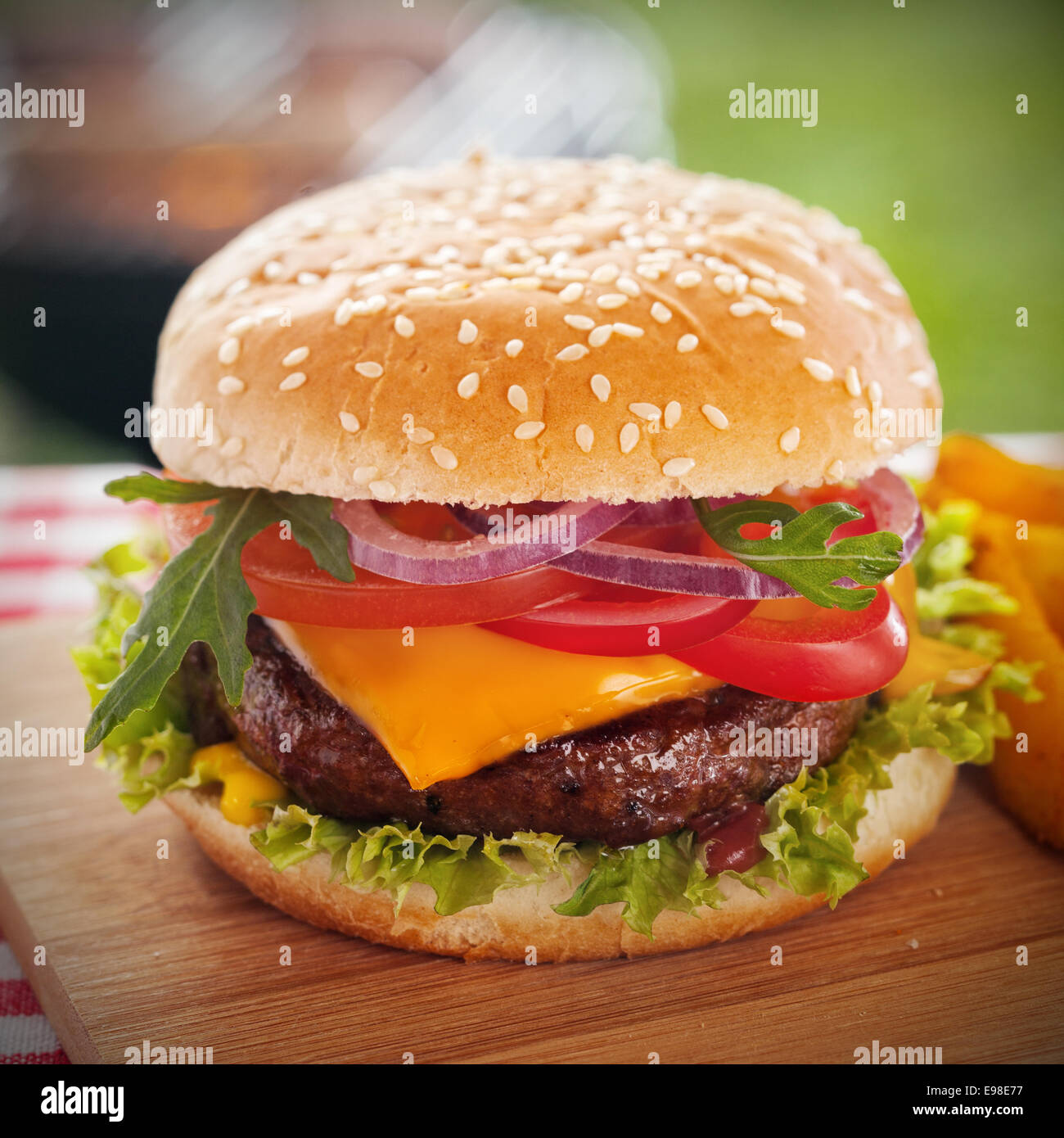 Tasty burger with melted cheese and a thick succulent ground beef patty garnished with lettuce, tomato, onion and rocket on a sesame bun standing on a picnic table in the garden Stock Photo
