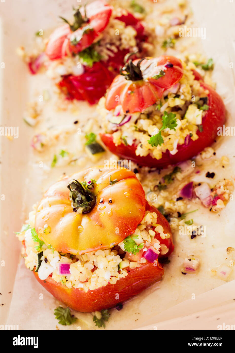 Ripe red baked tomato with savory corn and grain filling with chopped onion and herbs , high angle view of a row of three vegetables Stock Photo