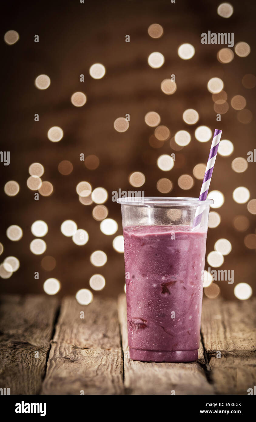 Energising berry smoothie in a plastic glass with a straw standing on a rustic wooden counter at a pub or diner with a background bokeh of sparkling lights Stock Photo