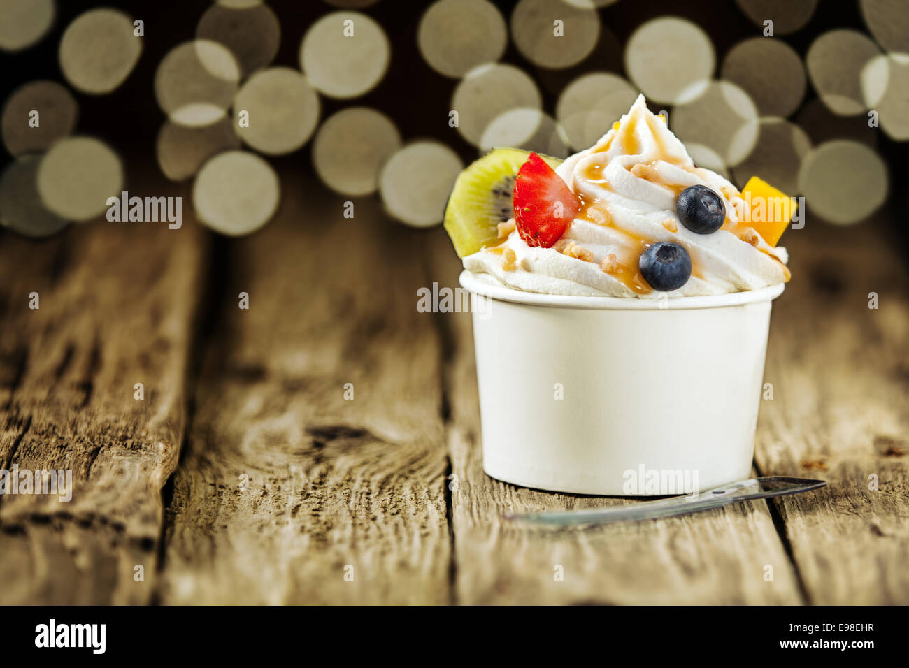 Creamy vanilla frozen yogurt topped with fresh tropical fruit served in a plastic takeaway tub with a disposable teaspoon on a wooden table with a background bokeh of party lights Stock Photo