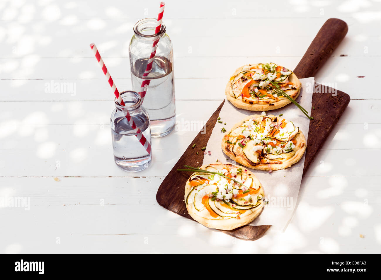 A trio of tasty mini vegetarian pizzas with eggplant, bell pepper, cheese and herbs served on a long wooden board with glass jars of fresh water Stock Photo