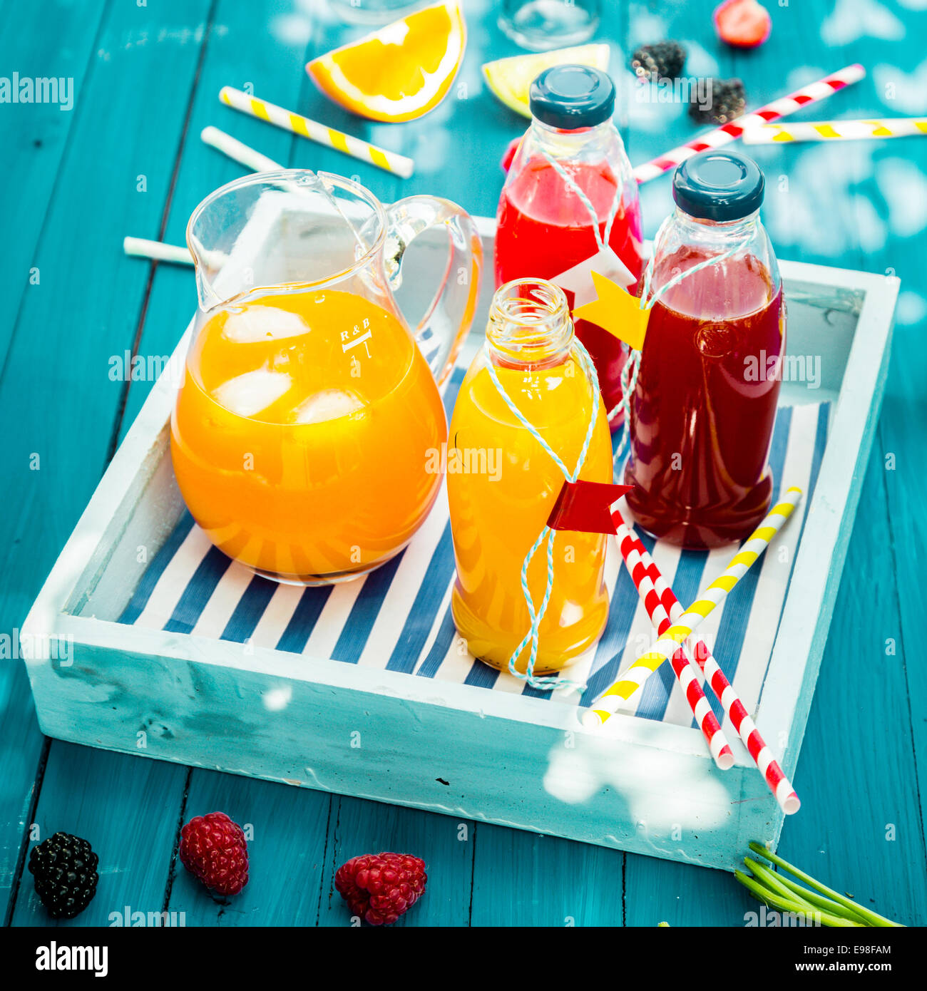 Bottles of freshly squeezed orange and berry juice standing on a wooden tray on a colorful turquoise blue picnic table in dappled summer sun Stock Photo