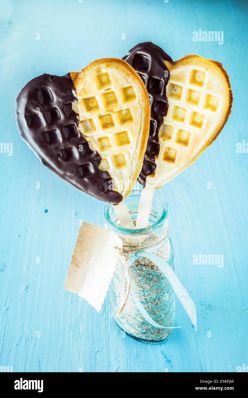 Two romantic heart shaped golden waffles dipped in chocolate symbolic of love on a stick in a glass jar with gift tag for a sweetheart on a special occasion or celebration Stock Photo