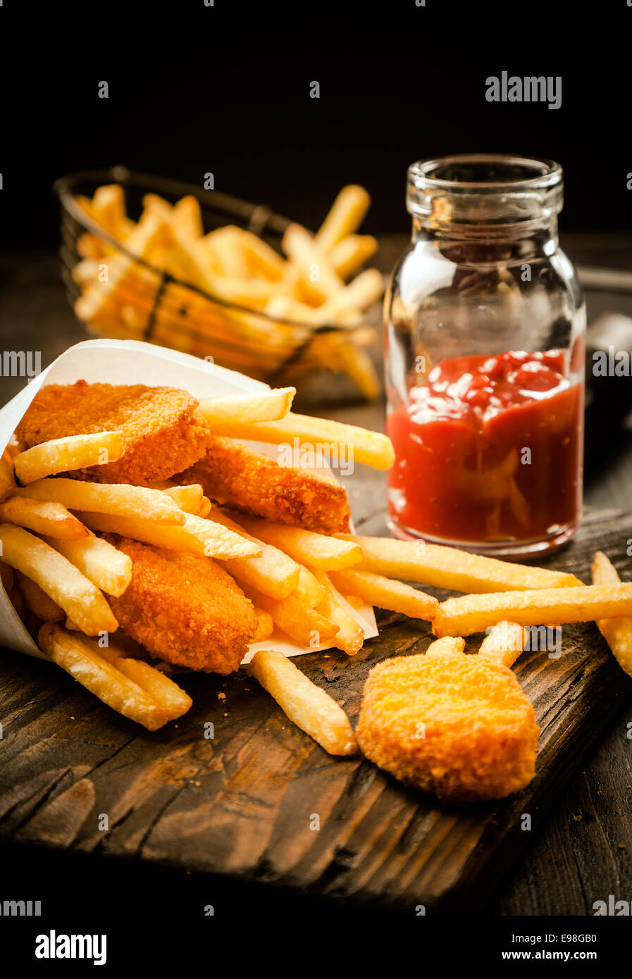 Fried golden crumbed fish fillets and French fries served in a paper cone with a tomato based sauce or dip on an old grunge wooden table in a fish shop Stock Photo