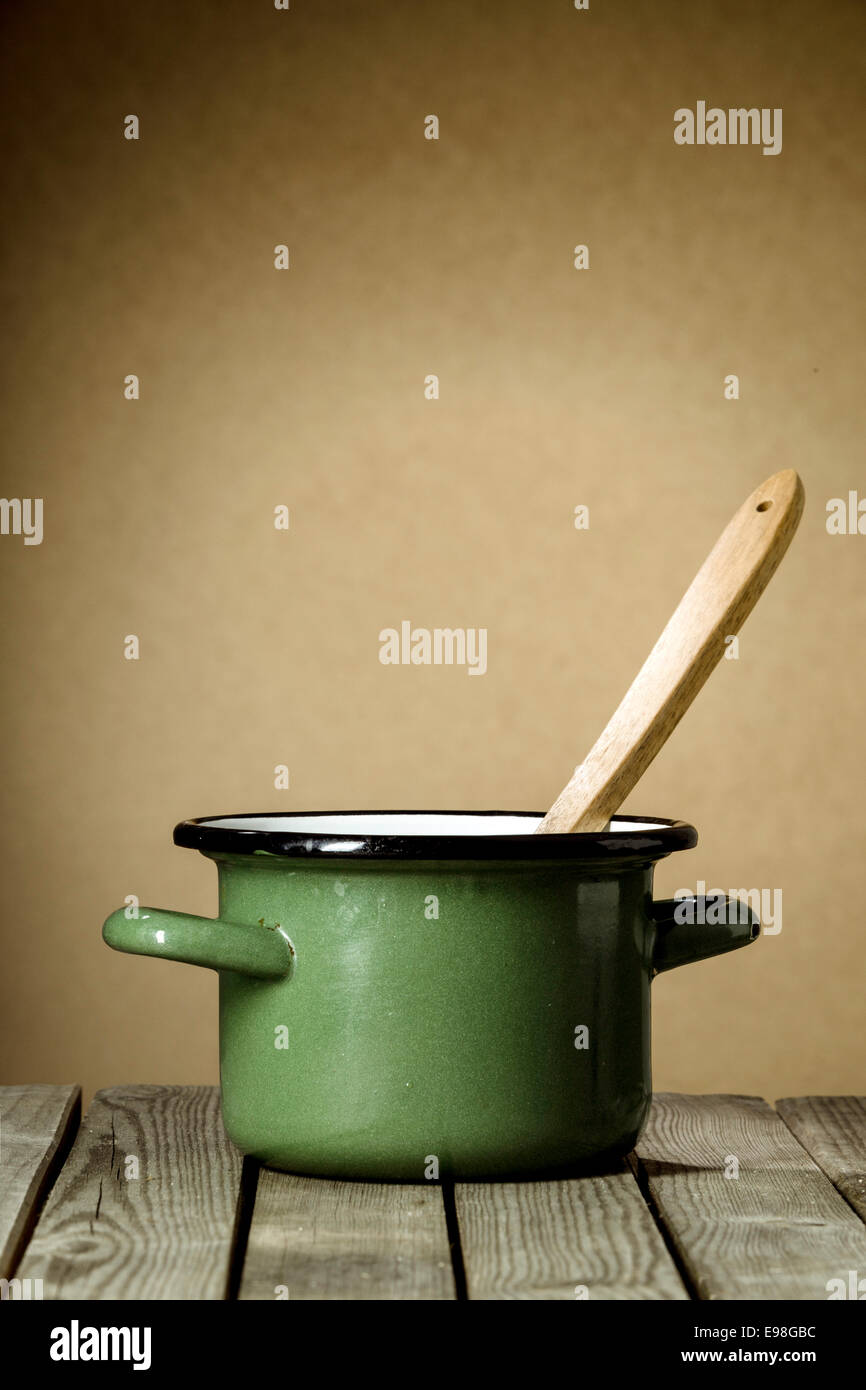 Rustic green enameled cooking pot with a wooden ladle inside standing on a wooden kitchen table against a brown wall with copyspace Stock Photo