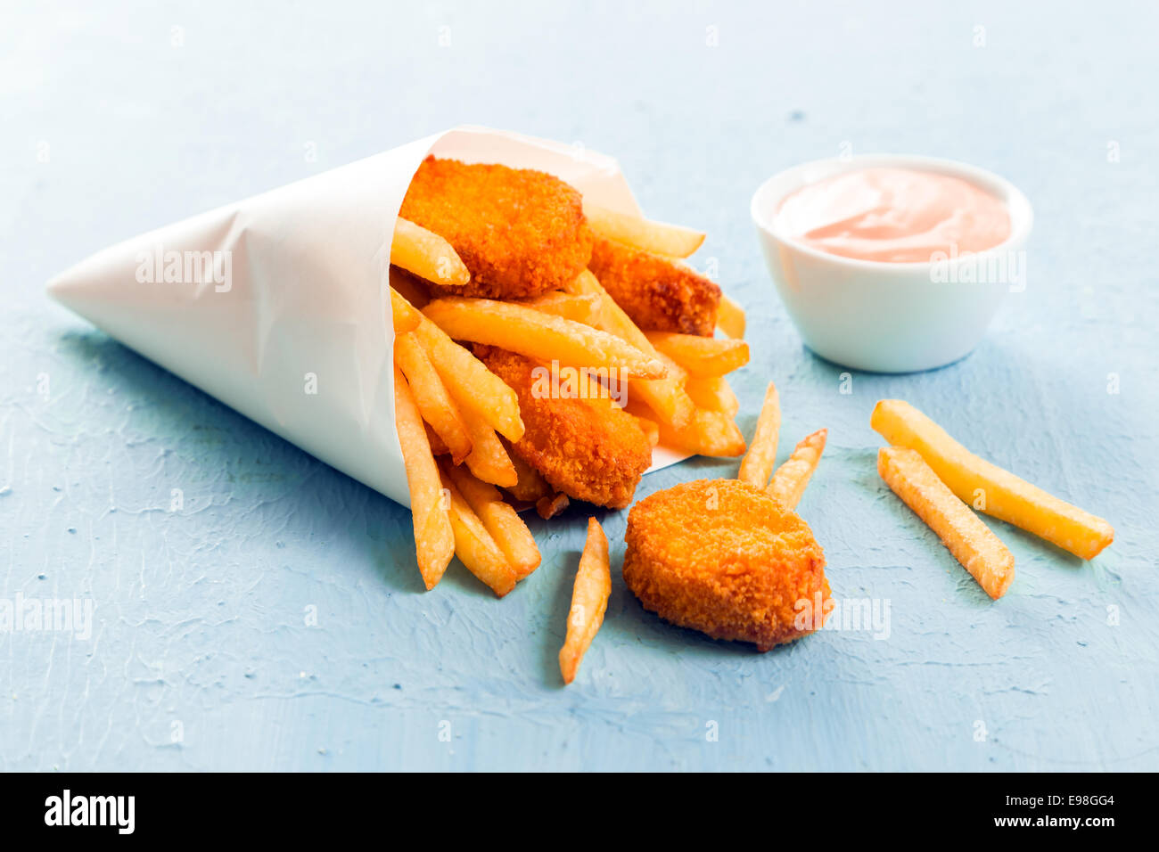 Golden fish nuggets with fried potato chips or French fries spilling out of a takeaway paper cone onto a blue surface with a small bowl of mayo dip Stock Photo