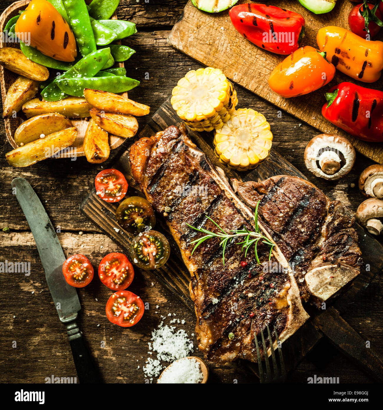 View from above of a delicious grilled porterhouse steak with assorted roast vegetables including tomato, peppers, mushrooms, corn, mangetout, and potato wedges Stock Photo