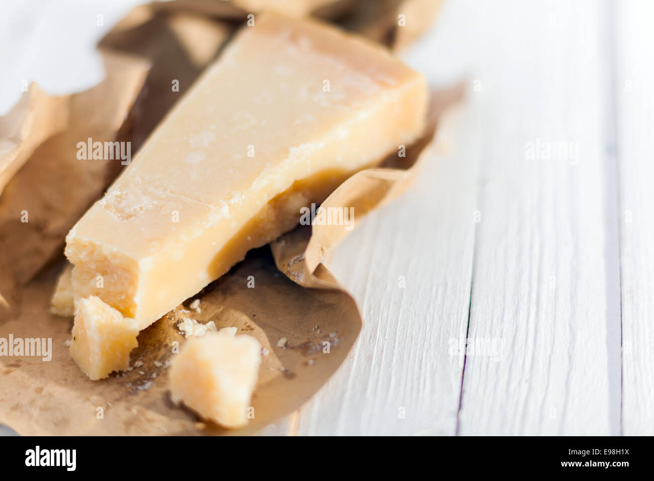Wedge of hardy crumbly matured Italian Parmigiano-Reggiano cheese, or Parmesan, in a torn brown paper wrapper on a white wooden Stock Photo