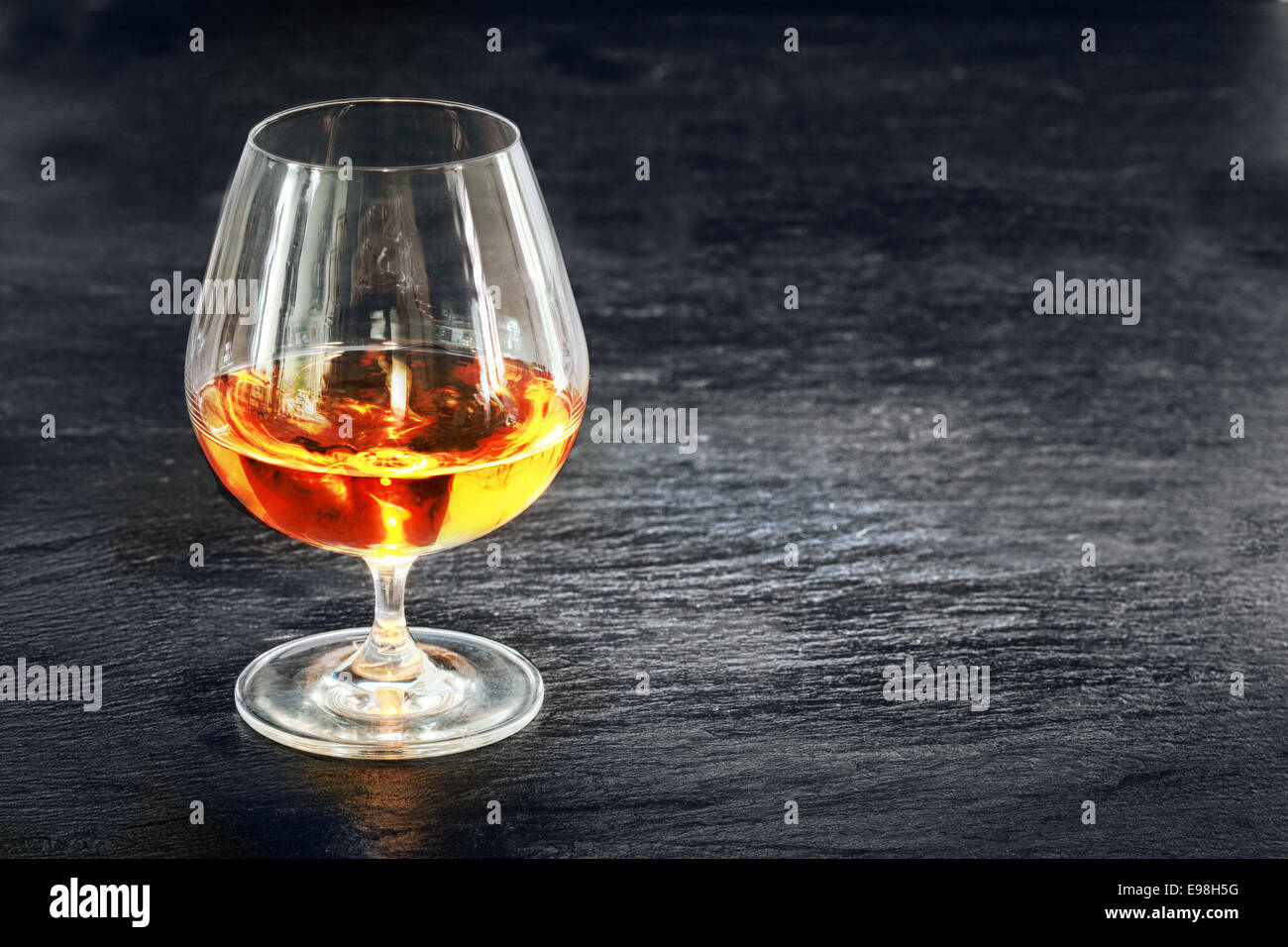 Elegant snifter with glowing golden cognac standing on a rustic dark bar counter with copyspace