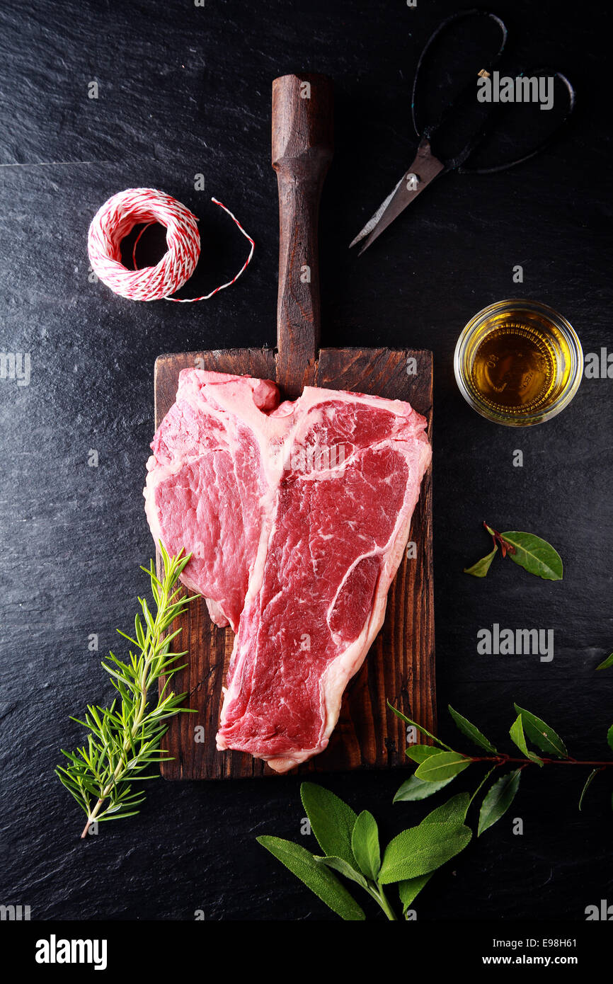Seasoning a t-bone steak for cooking with bay leaves, fresh rosemary, olive oil and string, overhead view on a wooden board on a rustic kitchen counter Stock Photo