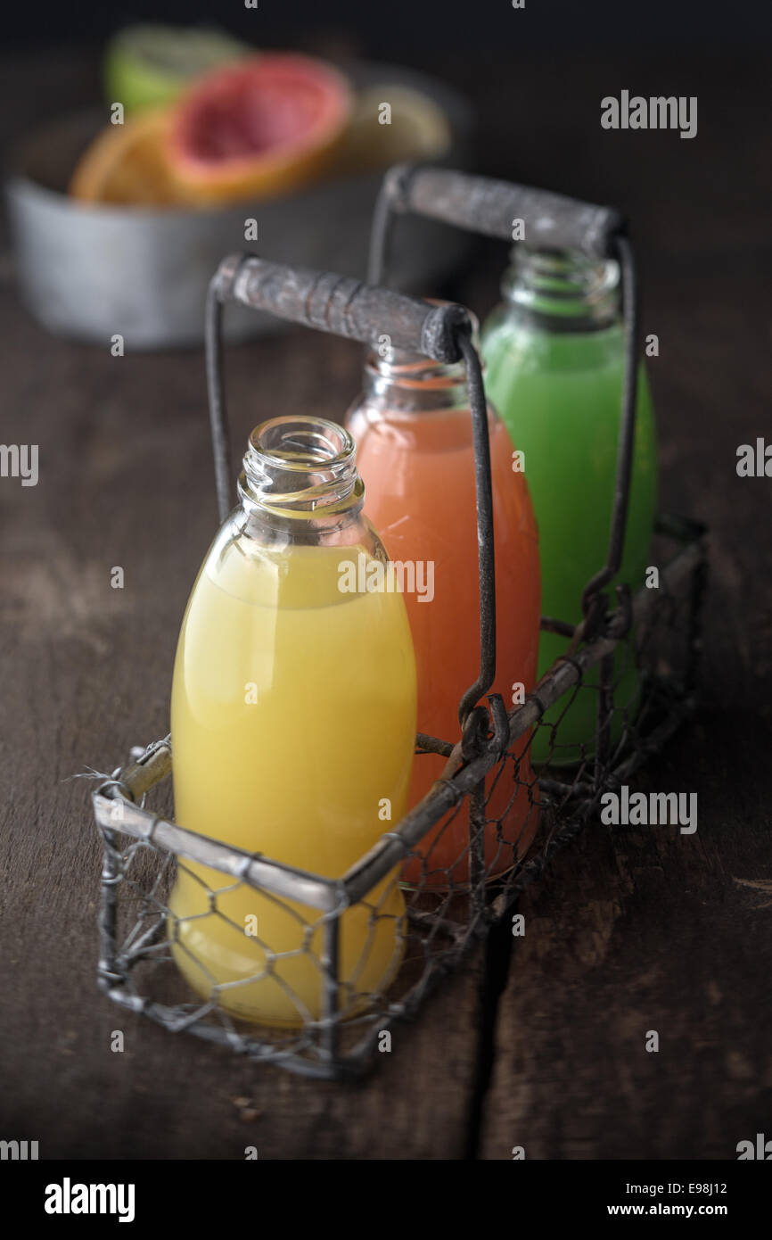 Glass bottles of assorted fresh fruit juice in an old metal and wire rack in a rustic kitchen with orange, grapefruit and lemon Stock Photo