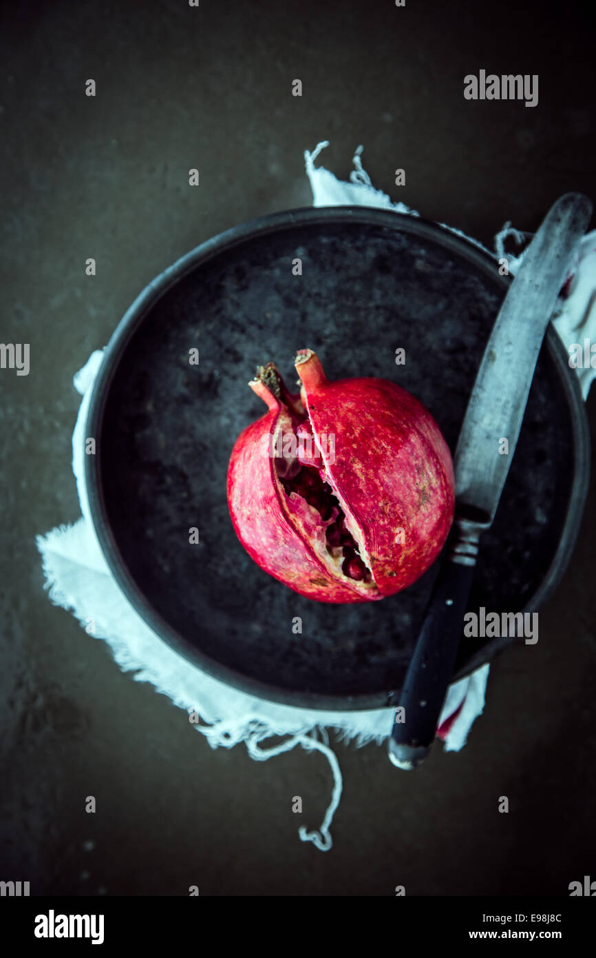 Splitting open a pomegranate with a large kitchen knife to access the juicy ripe red seeds, view from above on a plate against a dark background Stock Photo