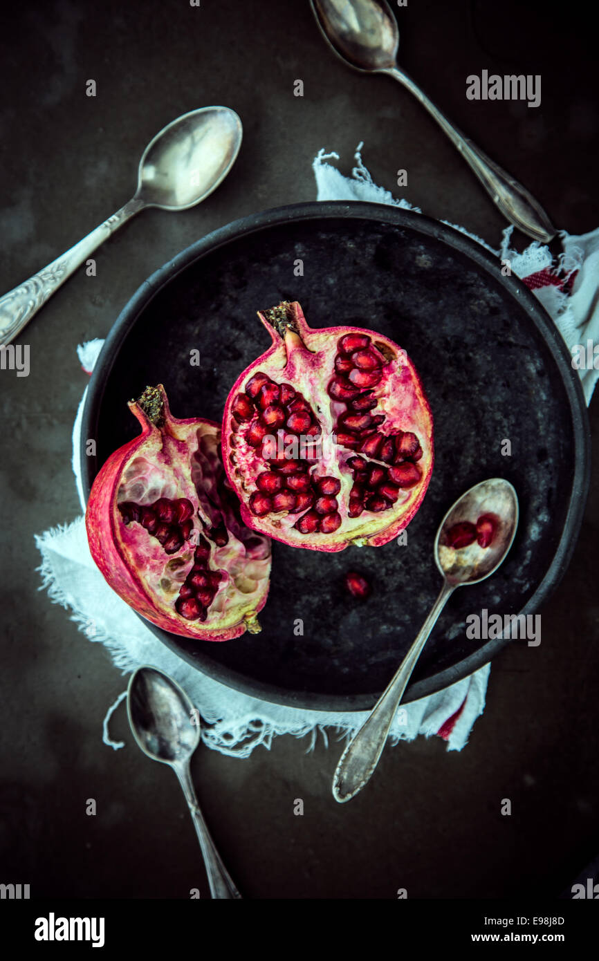 View from above of a fresh halved pomegranate on a plate showing the juicy ripe red sweet seeds with spoons for eating on a dark background Stock Photo