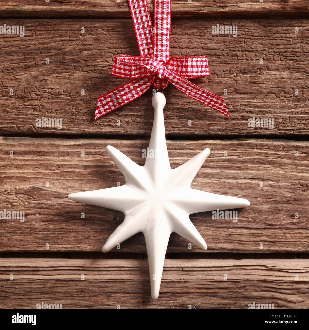 White Christmas star ornament hanging from a pretty red and white checked ribbon and bow on rustic textured wood in square format Stock Photo