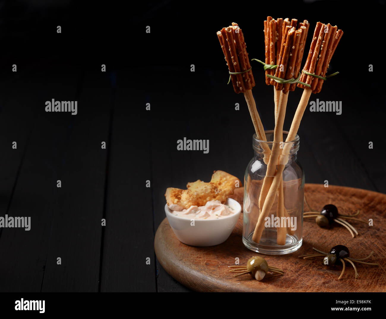 Witches broomsticks at a Halloween party made from bread sticks and pretzels standing in a glass jar on a table decorated with creepy spiders made from olives and spaghetti Stock Photo