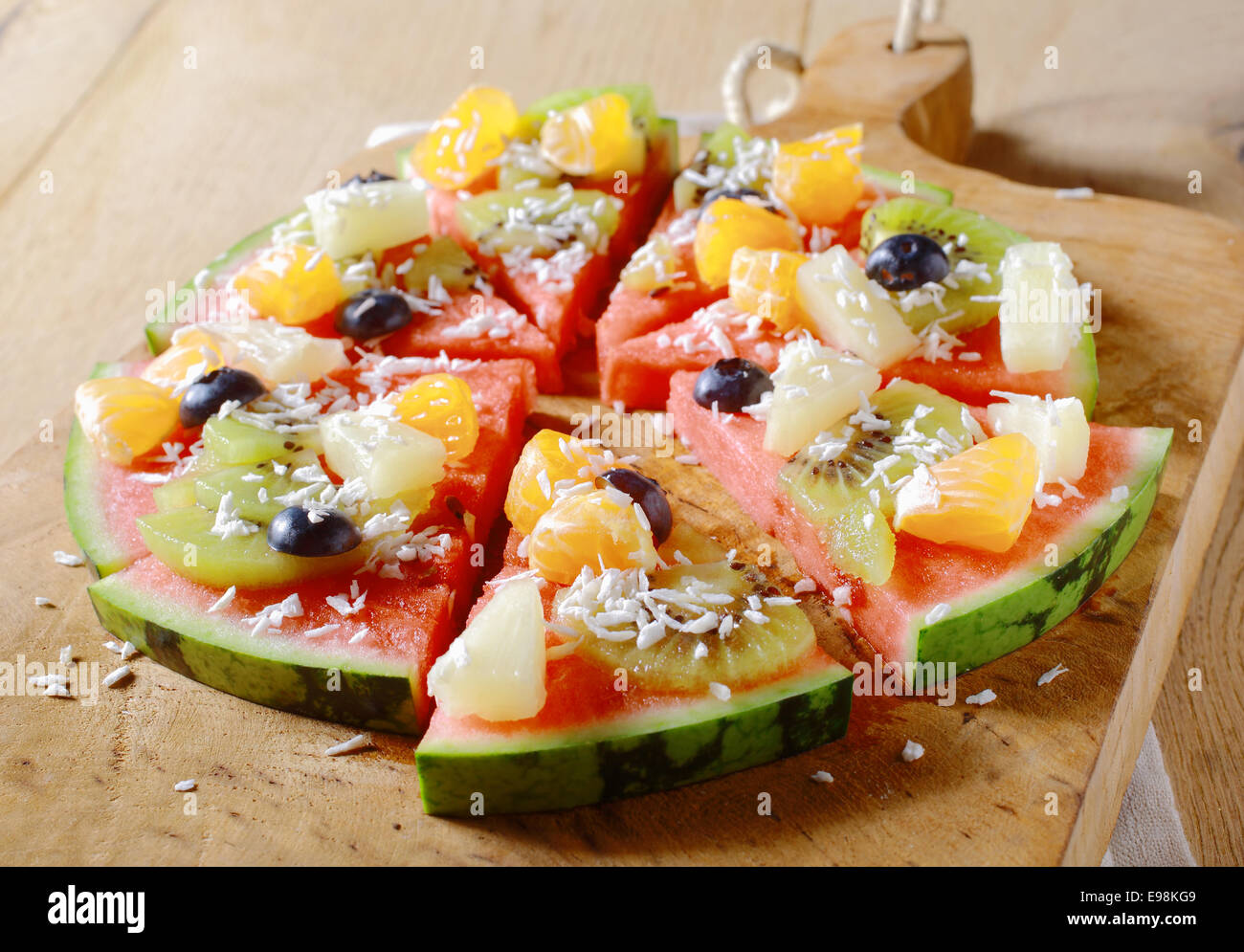 A tasty juicy fresh tropical fruit watermelon pizza topped with kiwifruit, blueberries, orange, pineapple, and topped with dried coconut, cut into segments on a wooden board for a nutritious dessert Stock Photo
