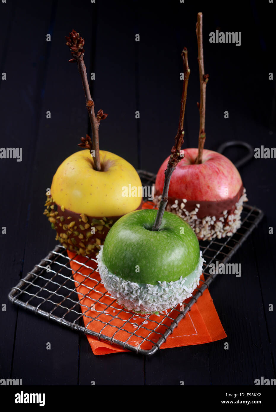 Three green, yellow and red candy coated apples dipped in chocolate, nuts and sprinkles for Halloween trick-or-treating treats standing cooling on a wire rack over a dark background Stock Photo