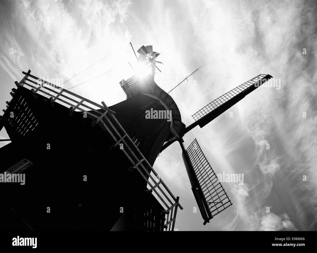 Monochrome image of a tilted windmill with it sails and balcony silhouetted against a cloudy sky by a sun flare over the roof. Stock Photo