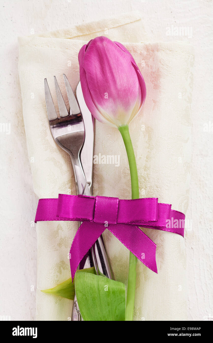 Napkin with tulip Flower. Dinner Table setting for two with a pink ribbon and a fork and a knife on wooden white background Stock Photo